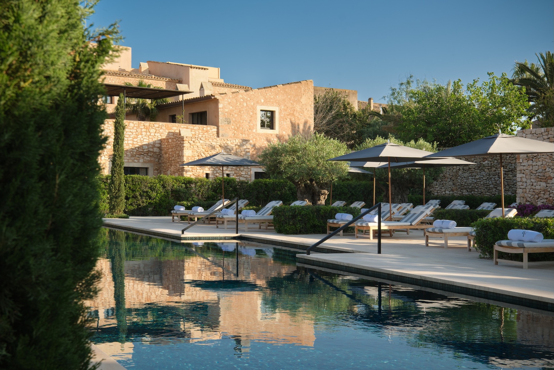 The pool at Can Ferrereta reflecting marès stone walls, parasols and a centenary olive tree, Santanyí | The Aficionados