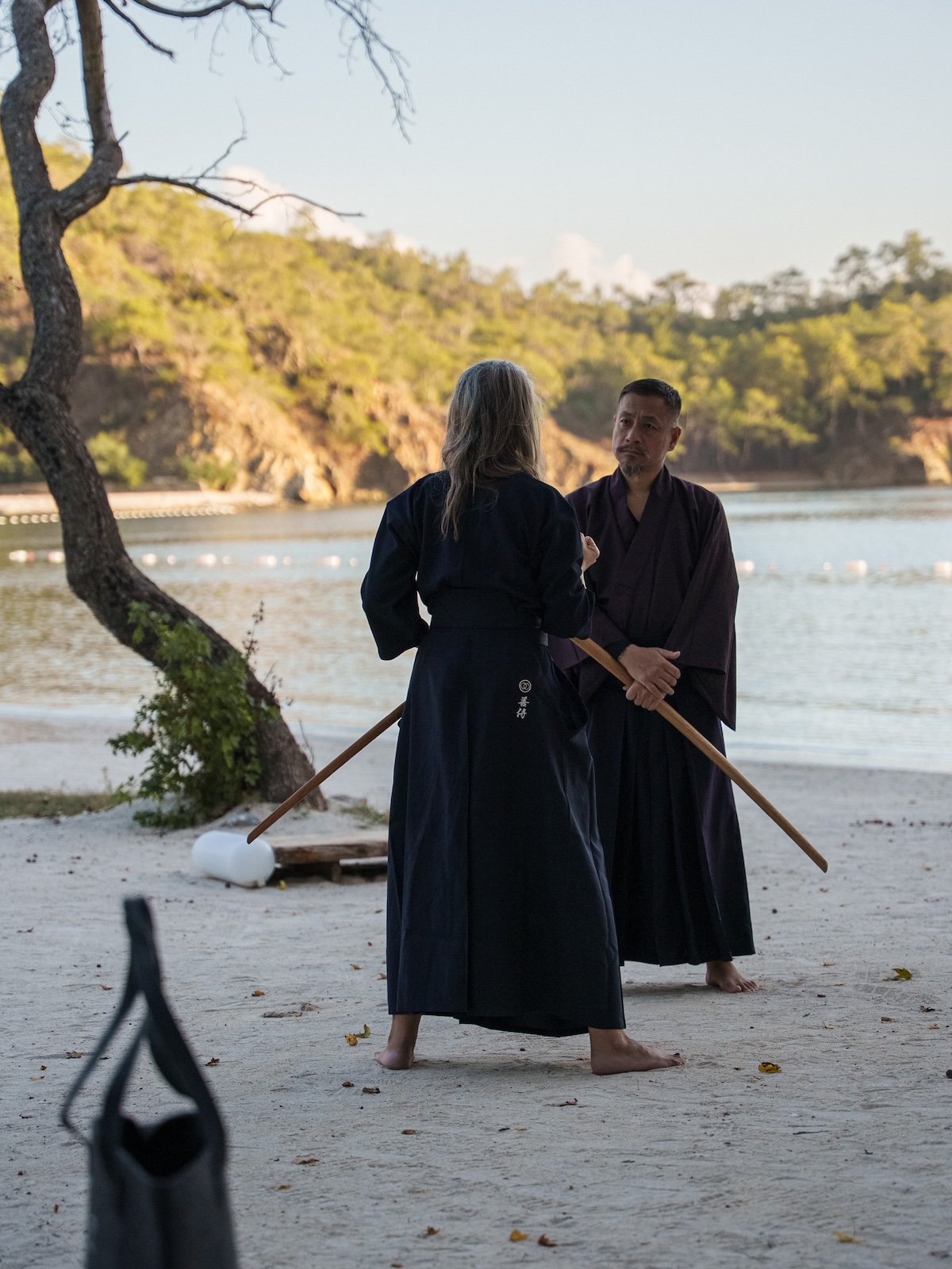 Two participants practise sword training as moving meditation with wooden bokken beside Sensei Zen Takai on the beach at AHAMA, Turkey.