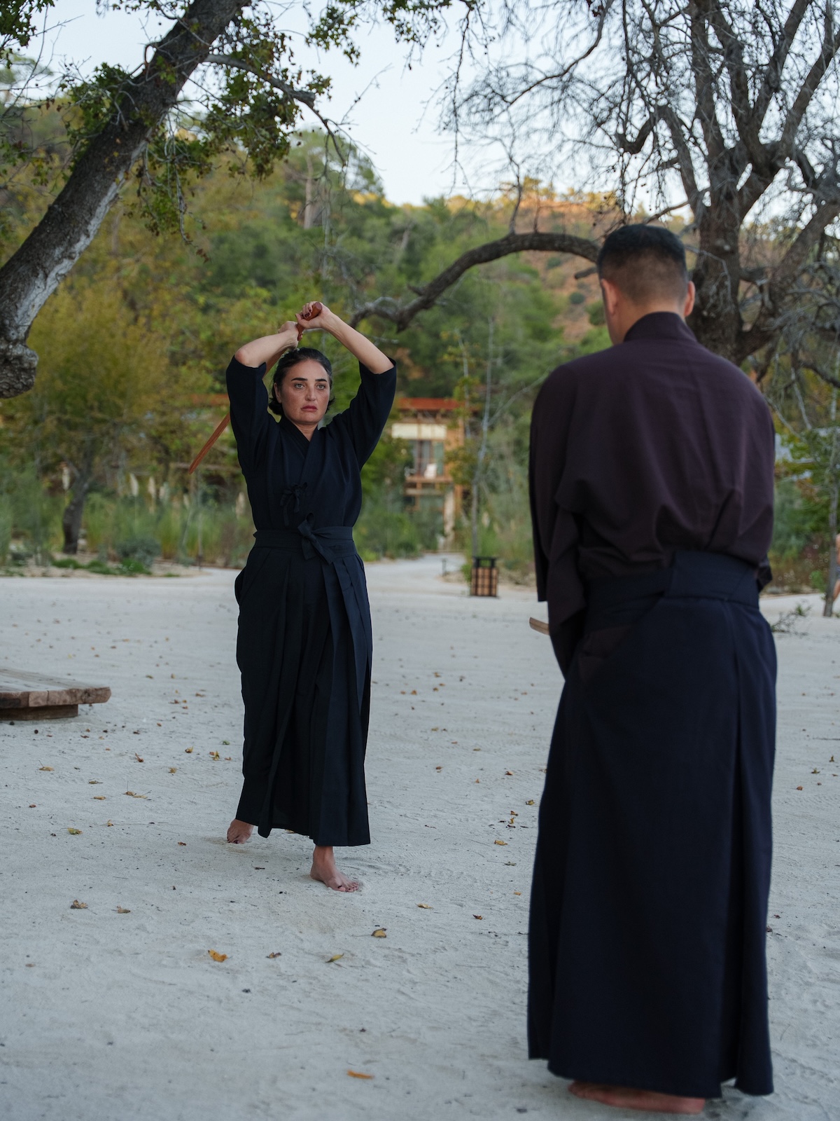 A retreat participant in black Samurai attire holds a barefoot training stance on the sand as Sensei Zen Takai observes at AHAMA, Turkey.