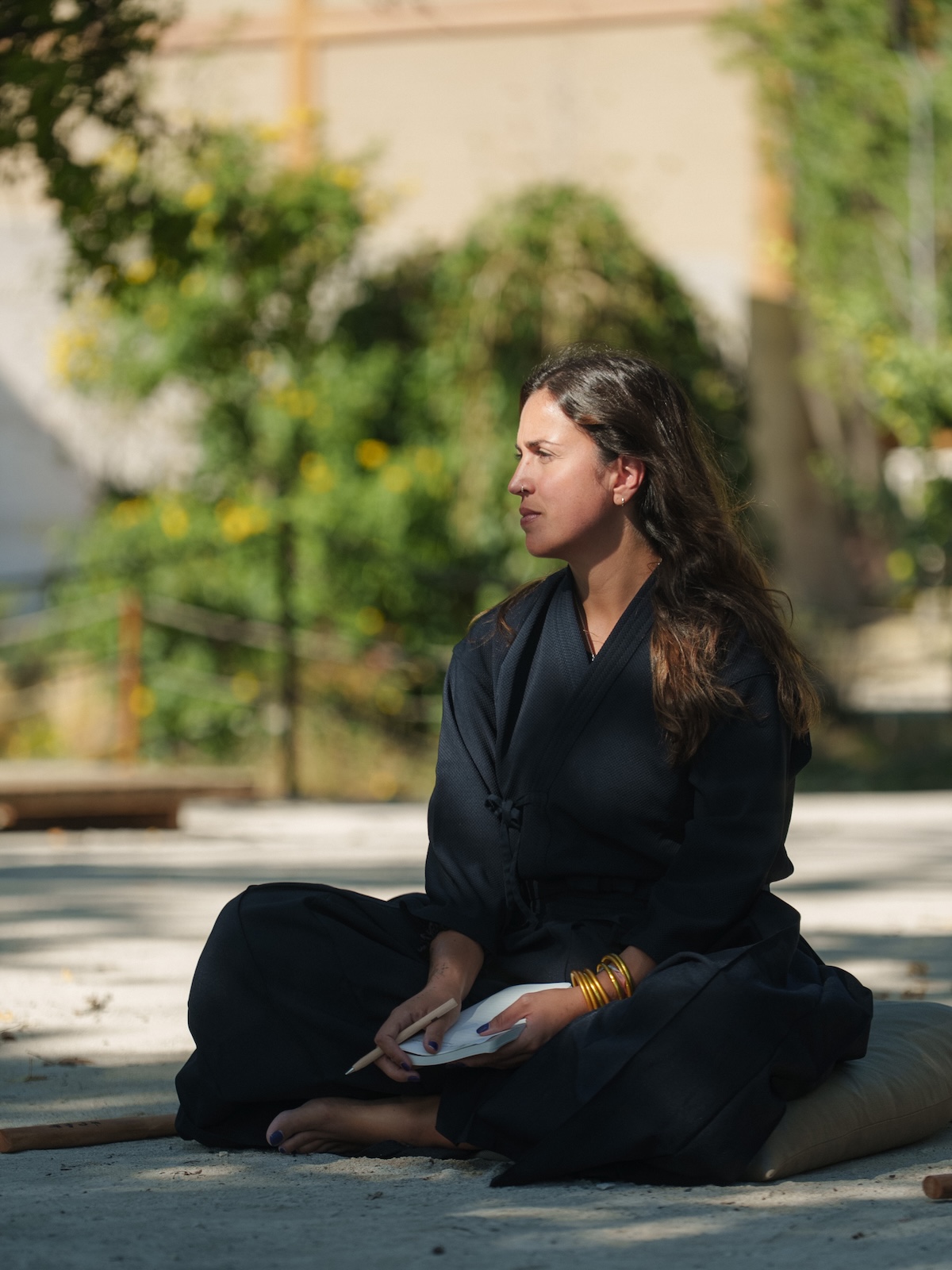 A woman in black Samurai robes sits in quiet meditation outdoors, holding calligraphy paper in the dappled morning light at the Zen Samurai retreat.