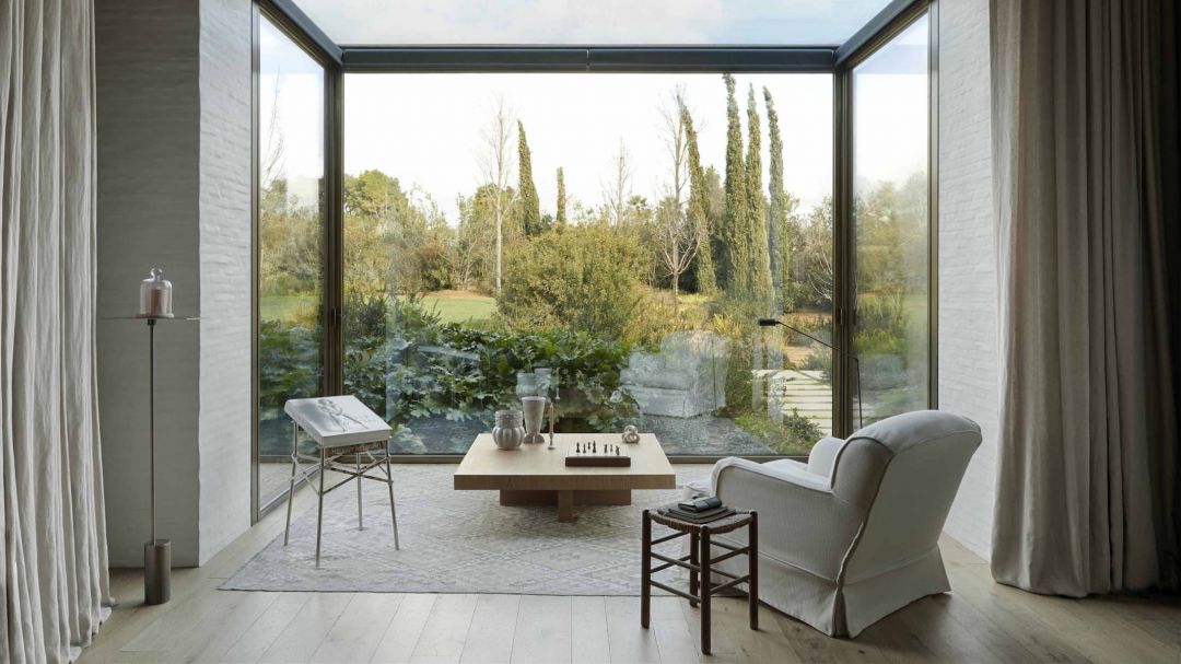 A glass-cornered sitting room framing a cypress garden at TATOÏ Club, near Athens, Greece, with a low table set for chess and a white slipper armchair on a pale rug.