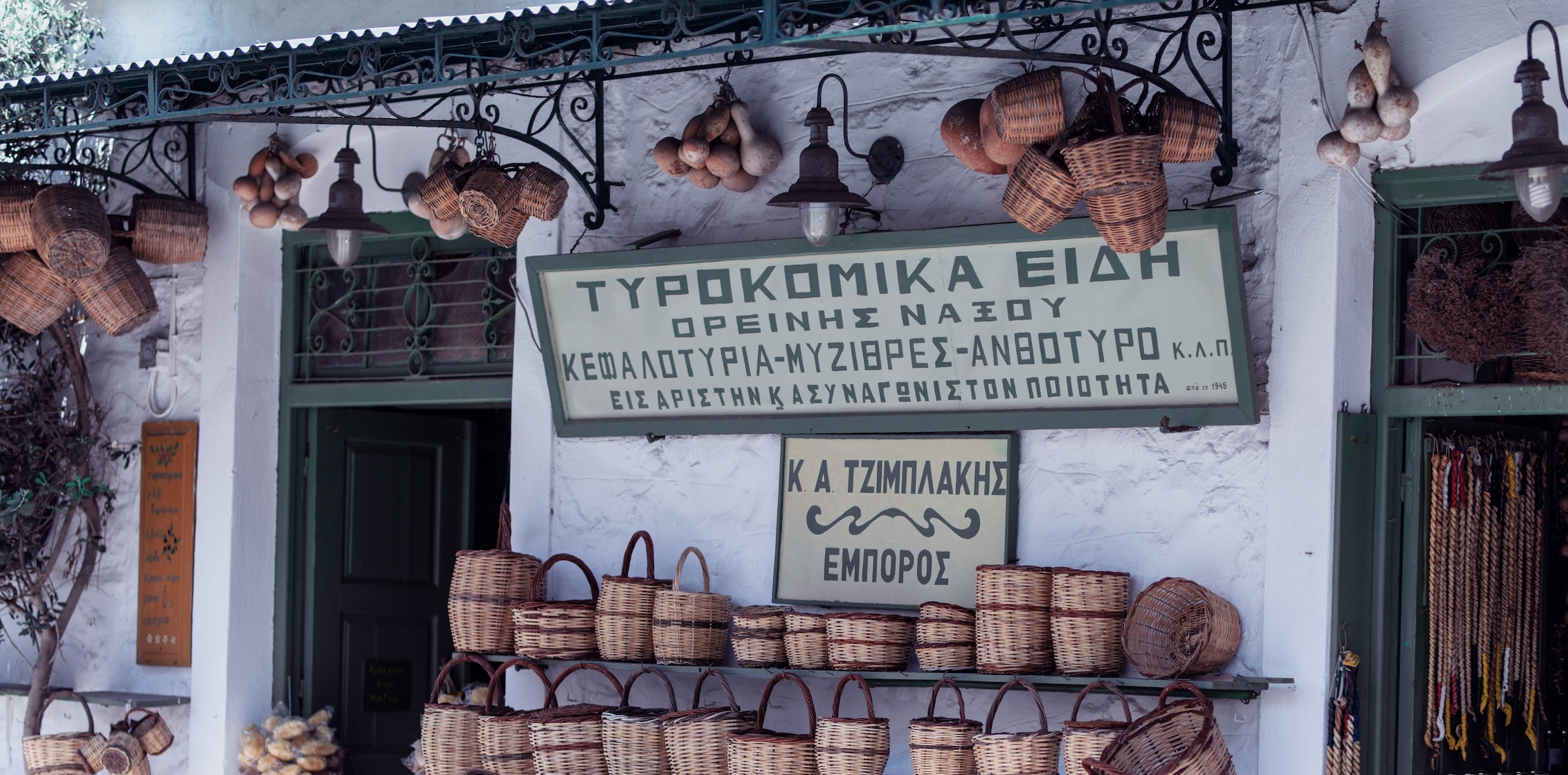 A traditional cheese and produce shop in the mountain villages of Naxos, wicker baskets and local Graviera beneath a hand-painted Greek sign.