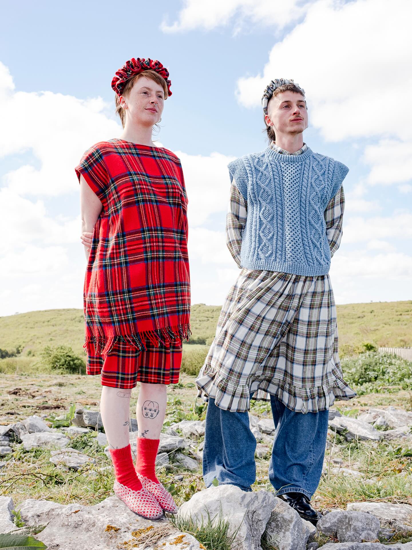 Two models on rocky ground: one in a fringed red tartan blanket dress and red woven shoes, the other in a pale blue Aran knit vest and checked ruffle-hem skirt over jeans.