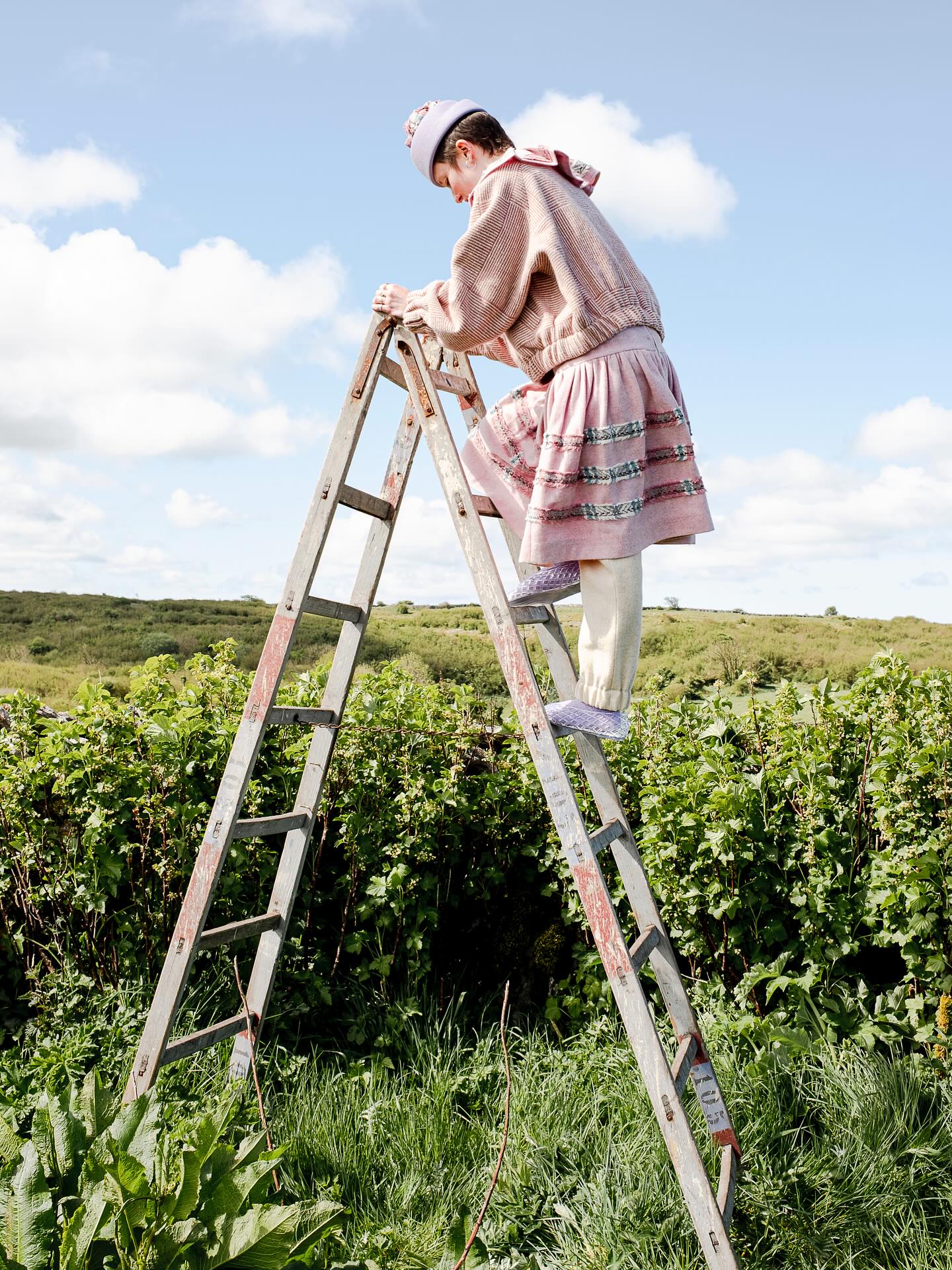 Model on a wooden stepladder among currant bushes, wearing a blush ribbed-knit bomber, a striped skirt and a lilac hat.