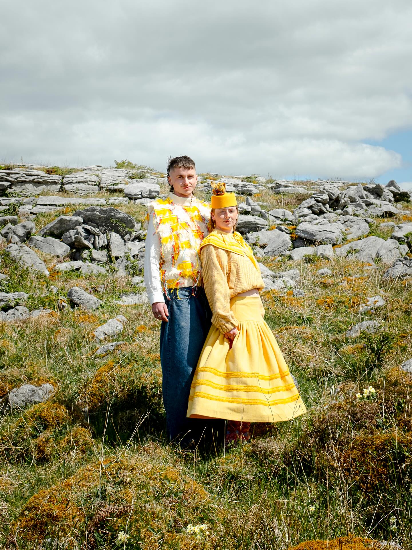 Couple on the Burren dressed head to toe in yellow: a fringed, tasselled tabard over a white shirt, beside a striped linen skirt, cropped knit and pillbox hat.