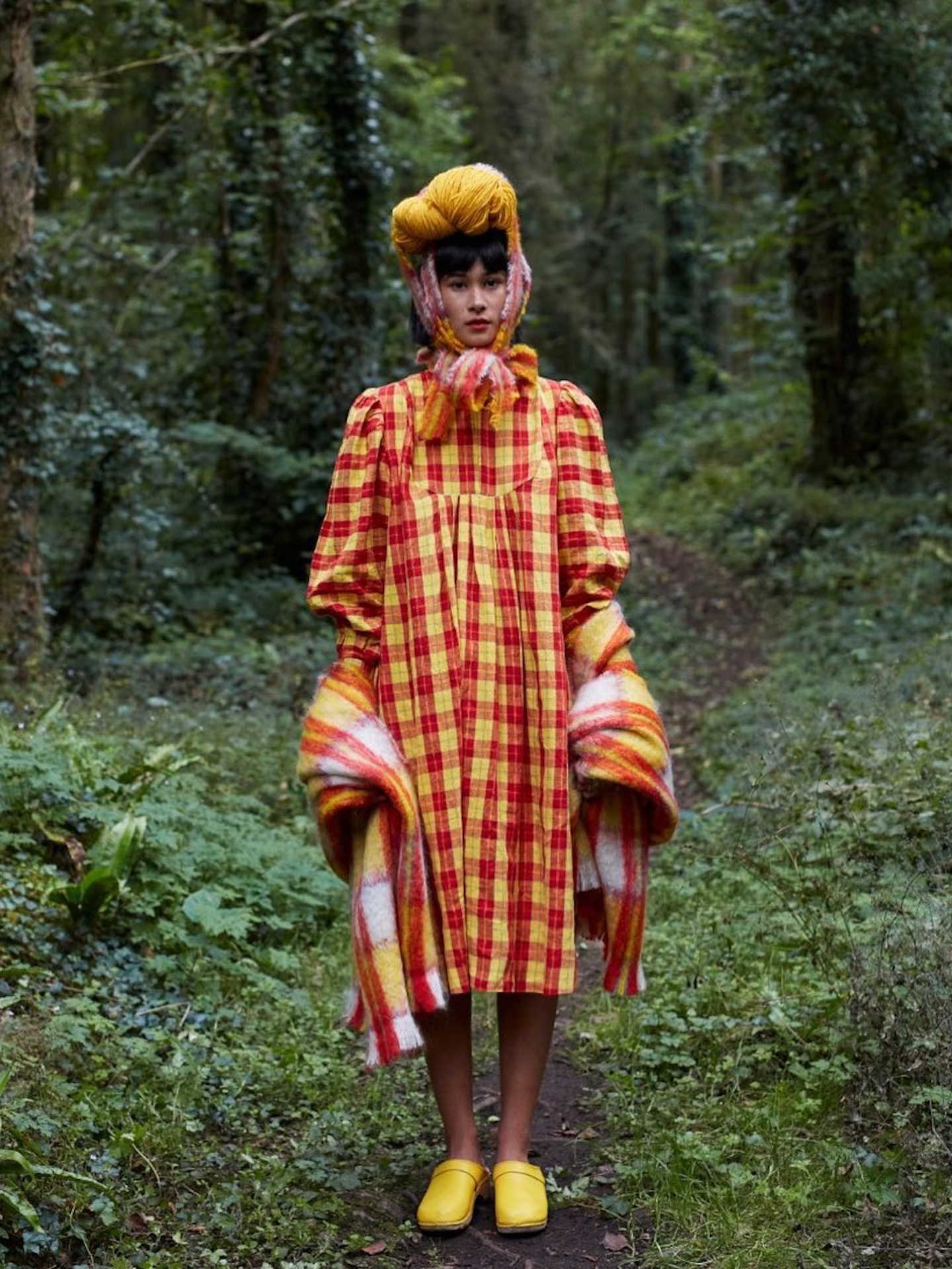 Model on a woodland path in a red and yellow checked smock dress and yellow wool turban, a striped mohair blanket draped over both arms.