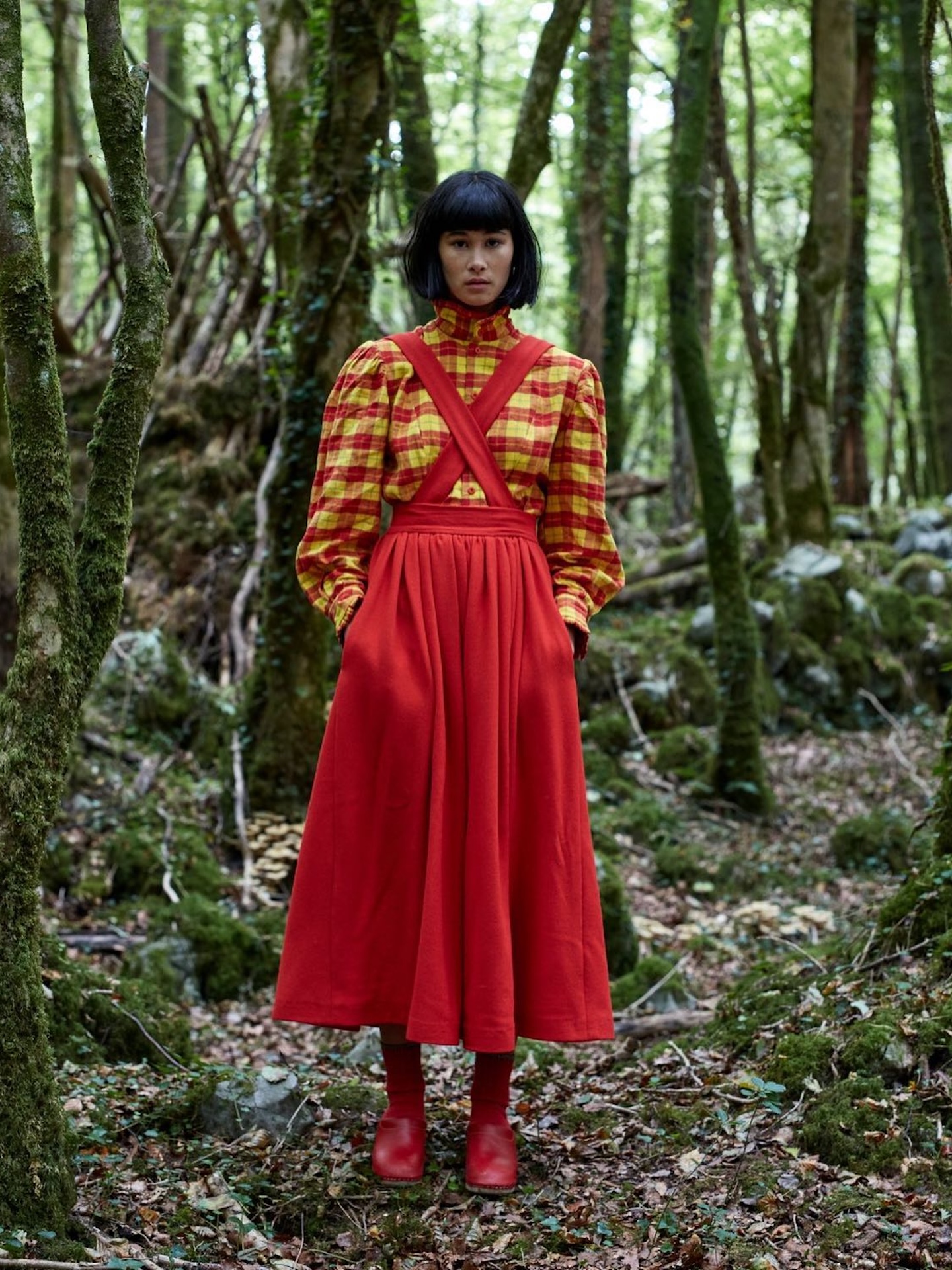 Model in mossy woodland in a red and yellow checked puff-sleeve blouse and red cross-strap pinafore skirt with red boots.