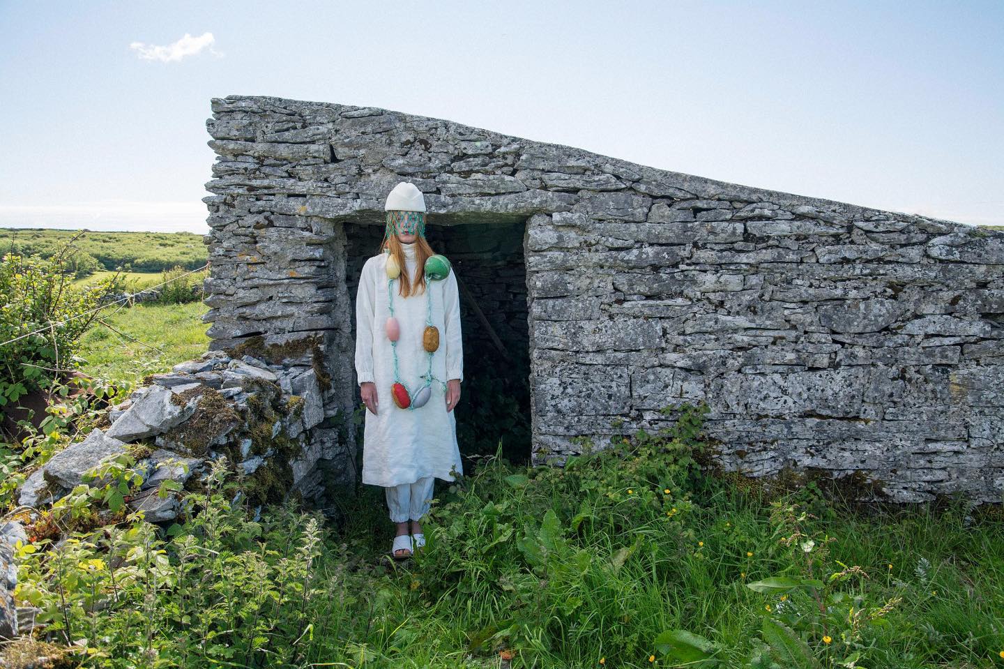 Model in a dry-stone doorway wearing a white linen tunic and trousers, a beaded veil and a necklace strung with colourful net-wrapped floats.