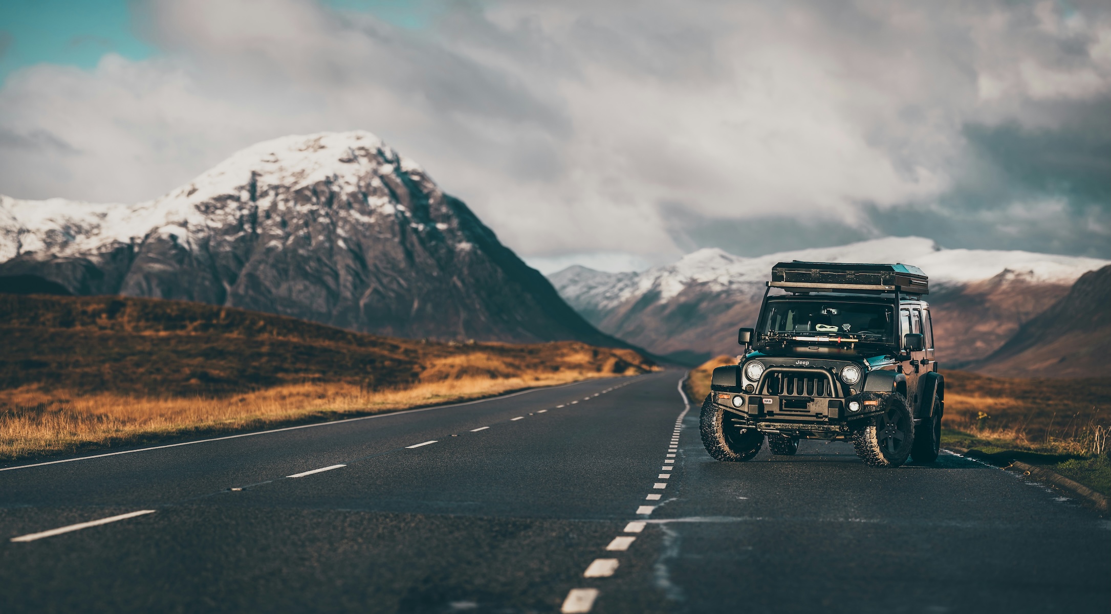 A rugged Jeep with a rooftop tent on an empty single-track road through Glencoe, snow-capped Highland mountains beyond, Scotland.