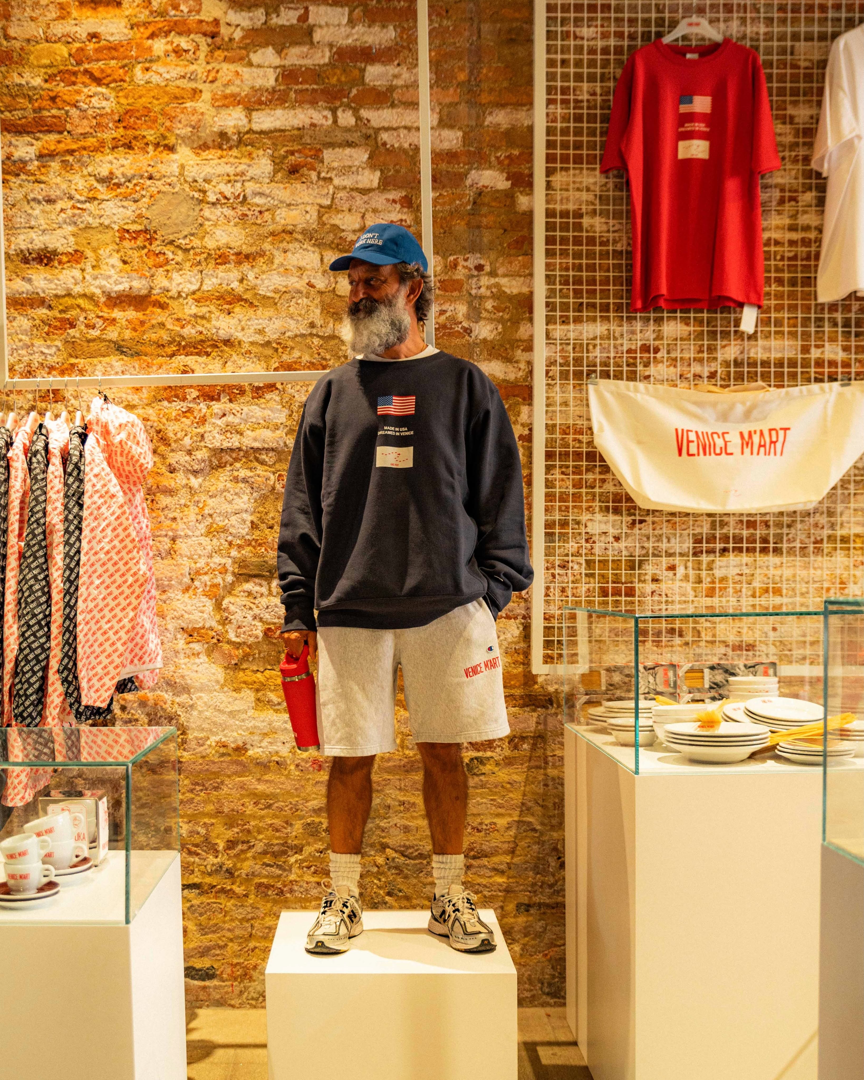 A man stands on a white plinth wearing a navy US-flag sweatshirt and grey Venice M'Art shorts, holding a red flask, beside a red flag tee and canvas Venice M'Art banner on an exposed brick wall.