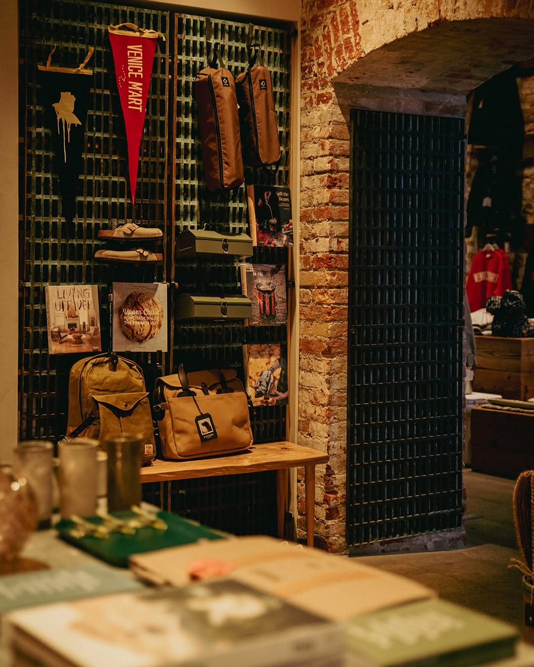 Corner display at Venice M'Art with a red Venice M'Art felt pennant, tan backpack and bags, Birkenstock sandals and design books arranged against dark tiled and brick walls.