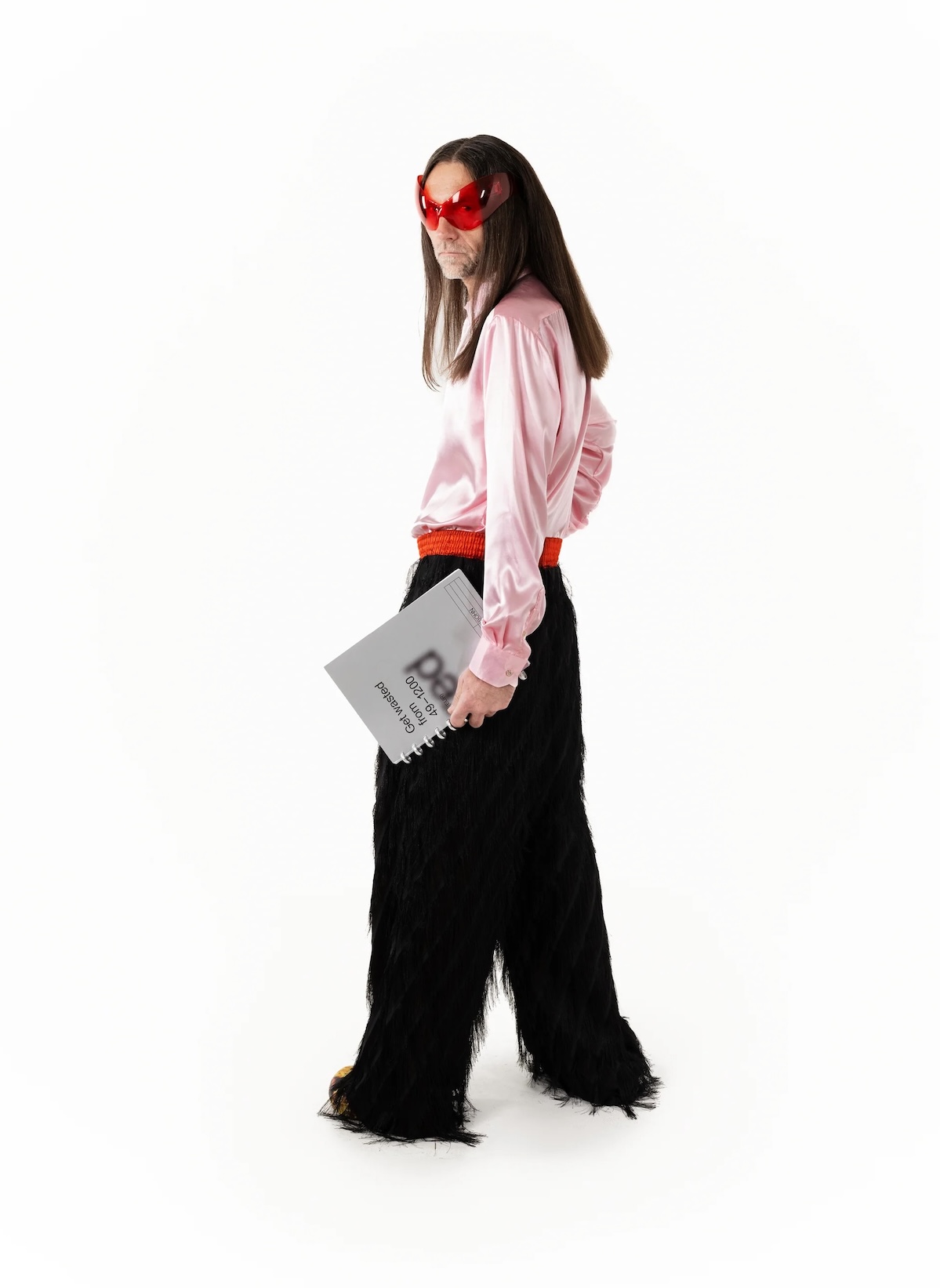 A figure in red heart-shaped sunglasses, pink satin shirt and shaggy black trousers holding an independent print magazine, in the spirit of Vienna's Softcover